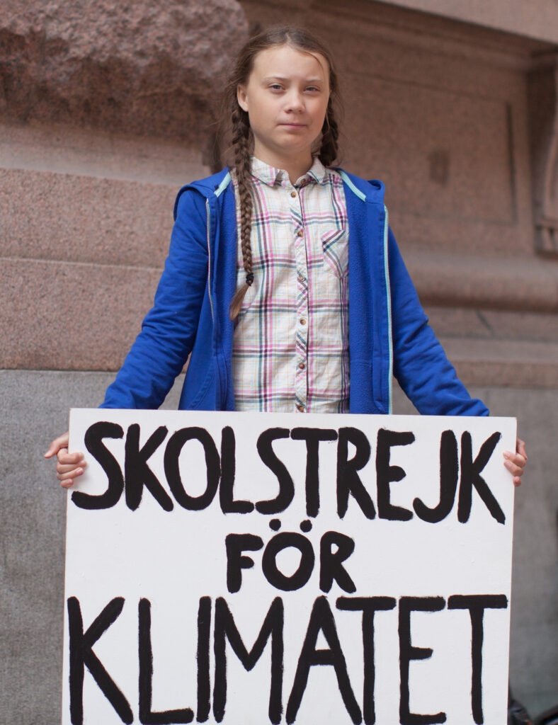 Die Klimaaktivistin Greta Thunberg spricht während einer Demonstration, erkennbar an ihrem ernsten Gesichtsausdruck und dem charakteristischen gelben Regenmantel.