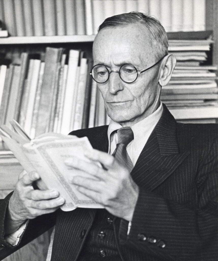 Hermann Hesse in seinem Garten in Montagnola, um 1950, mit Hut und Brille in nachdenklicher Pose sitzend.