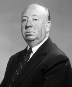 Alfred Hitchcock in typischer Pose auf einem Regiestuhl am Set, um 1960, mit ernstem Blick und Zigarre in der Hand.