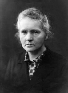 Marie Curie in ihrem Pariser Labor um 1912, sie sitzt an einem Tisch mit wissenschaftlichen Apparaturen und blickt nachdenklich auf ihre Instrumente.