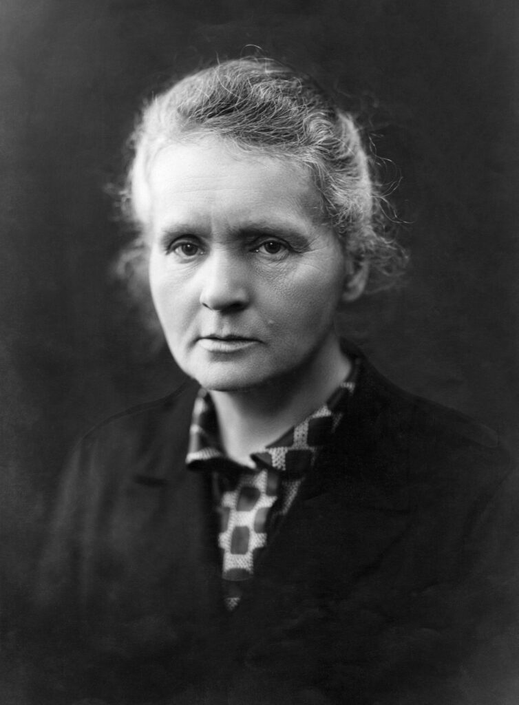 Marie Curie in ihrem Pariser Labor um 1912, sie sitzt an einem Tisch mit wissenschaftlichen Apparaturen und blickt nachdenklich auf ihre Instrumente.