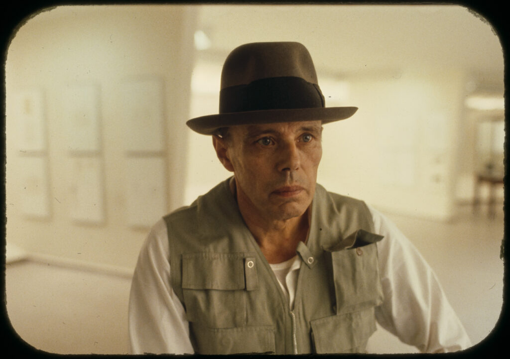 Joseph Beuys mit seinem charakteristischen Filzhut und einer Anglerweste während einer Aktion, sein Blick ist intensiv und konzentriert.