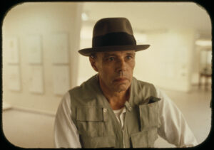 Joseph Beuys mit seinem charakteristischen Filzhut und einer Anglerweste während einer Aktion, sein Blick ist intensiv und konzentriert.