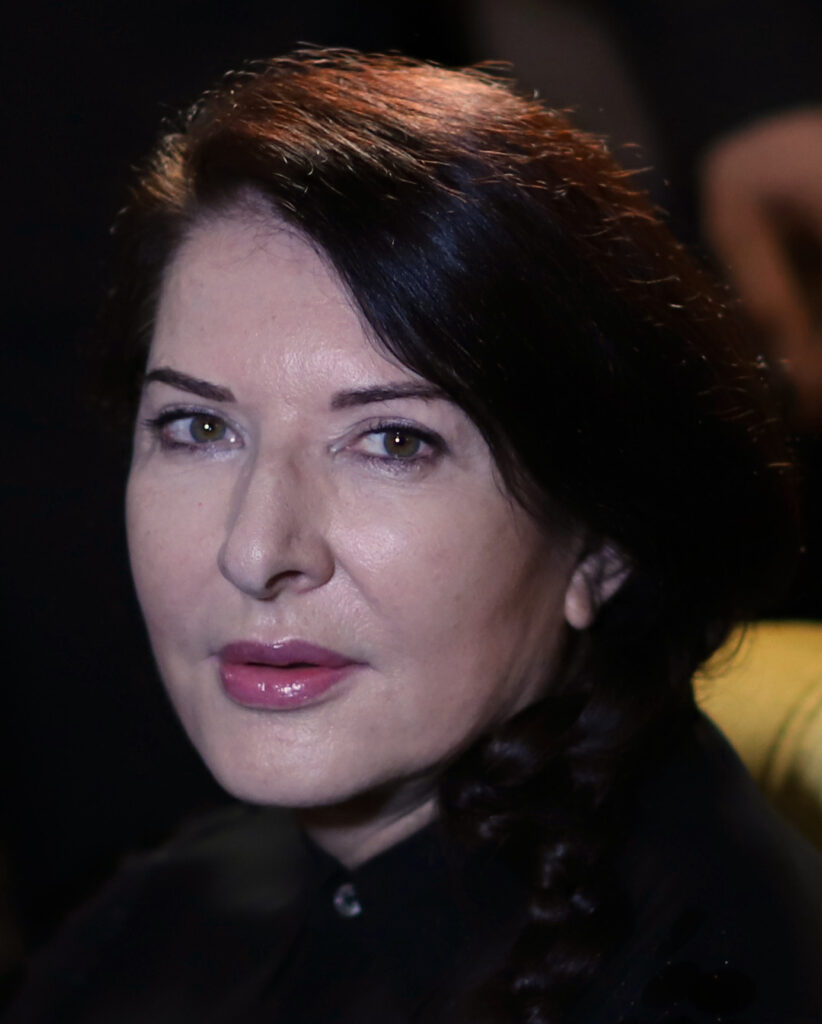 Marina Abramović während ihrer Performance 'The Artist is Present' im MoMA 2010, sie blickt mit ruhiger Intensität direkt in die Kamera.