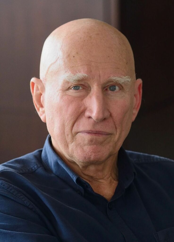 Der Fotograf Sebastião Salgado bei einer Ausstellungseröffnung, in schwarzweiß gehalten, mit einer seiner großformatigen Fotografien im Hintergrund.