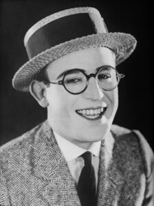 Der Stummfilmkomiker Harold Lloyd hängt am Zeiger einer riesigen Uhr an einer Hochhausfassade, eine ikonische Szene aus seinem Film Safety Last! (1923).