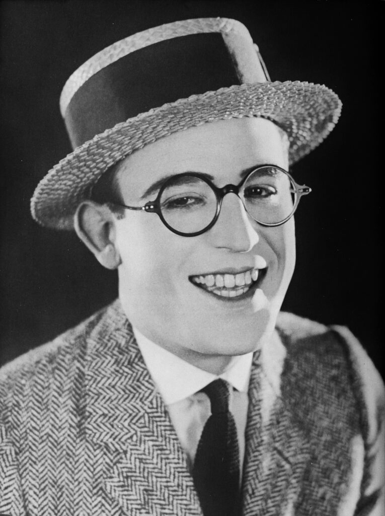 Der Stummfilmkomiker Harold Lloyd hängt am Zeiger einer riesigen Uhr an einer Hochhausfassade, eine ikonische Szene aus seinem Film Safety Last! (1923).